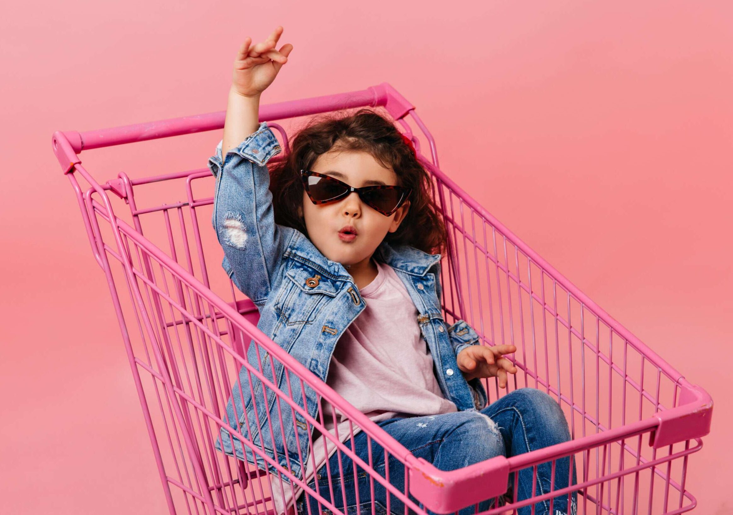 Funny child in sunglasses sitting in shopping cart. Studio shot of happy little girl in denim jacket.